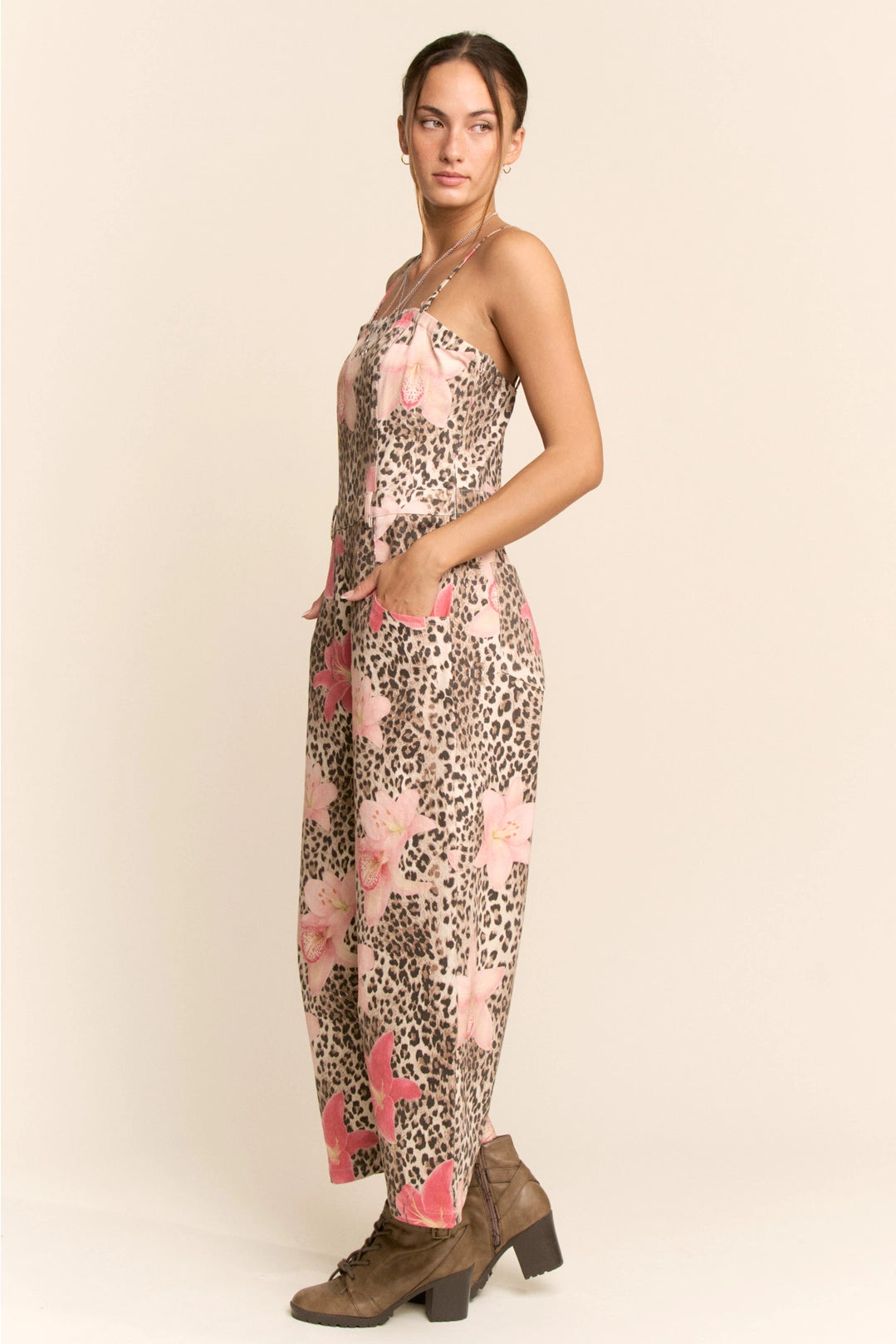 A woman modeling a sleeveless maxi dress with a pink and brown leopard print and large pink floral motifs, paired with brown heeled boots, against a solid beige background.