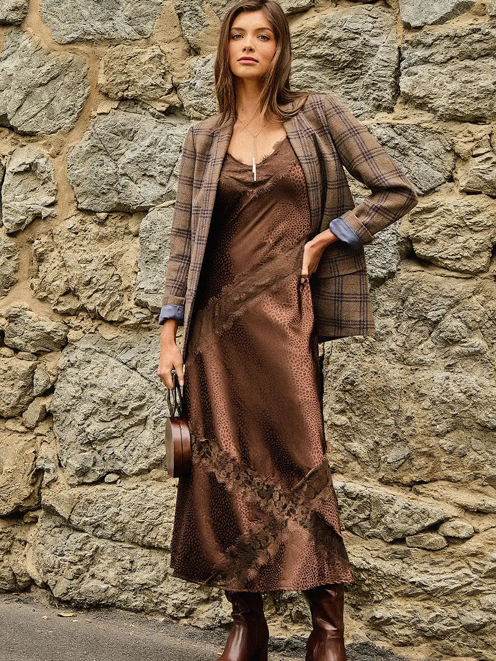 A woman stands against a rustic stone wall wearing a brown satin slip dress with a lace overlay, styled with a brown plaid blazer, brown boots, and holding a small brown handbag.