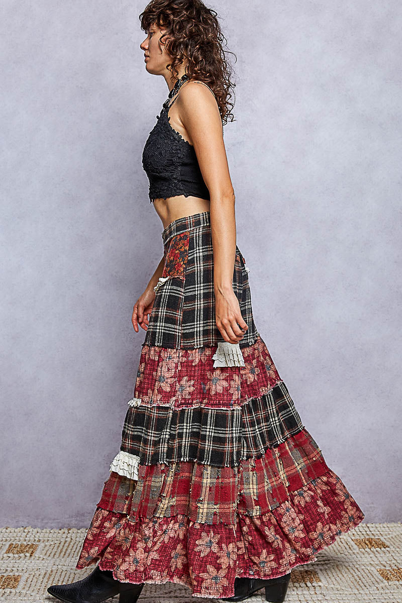 A woman in profile wearing a long, tiered patchwork maxi skirt with red, black, and beige plaid and floral patterns, paired with a black lace halter crop top, standing against a textured gray studio backdrop.