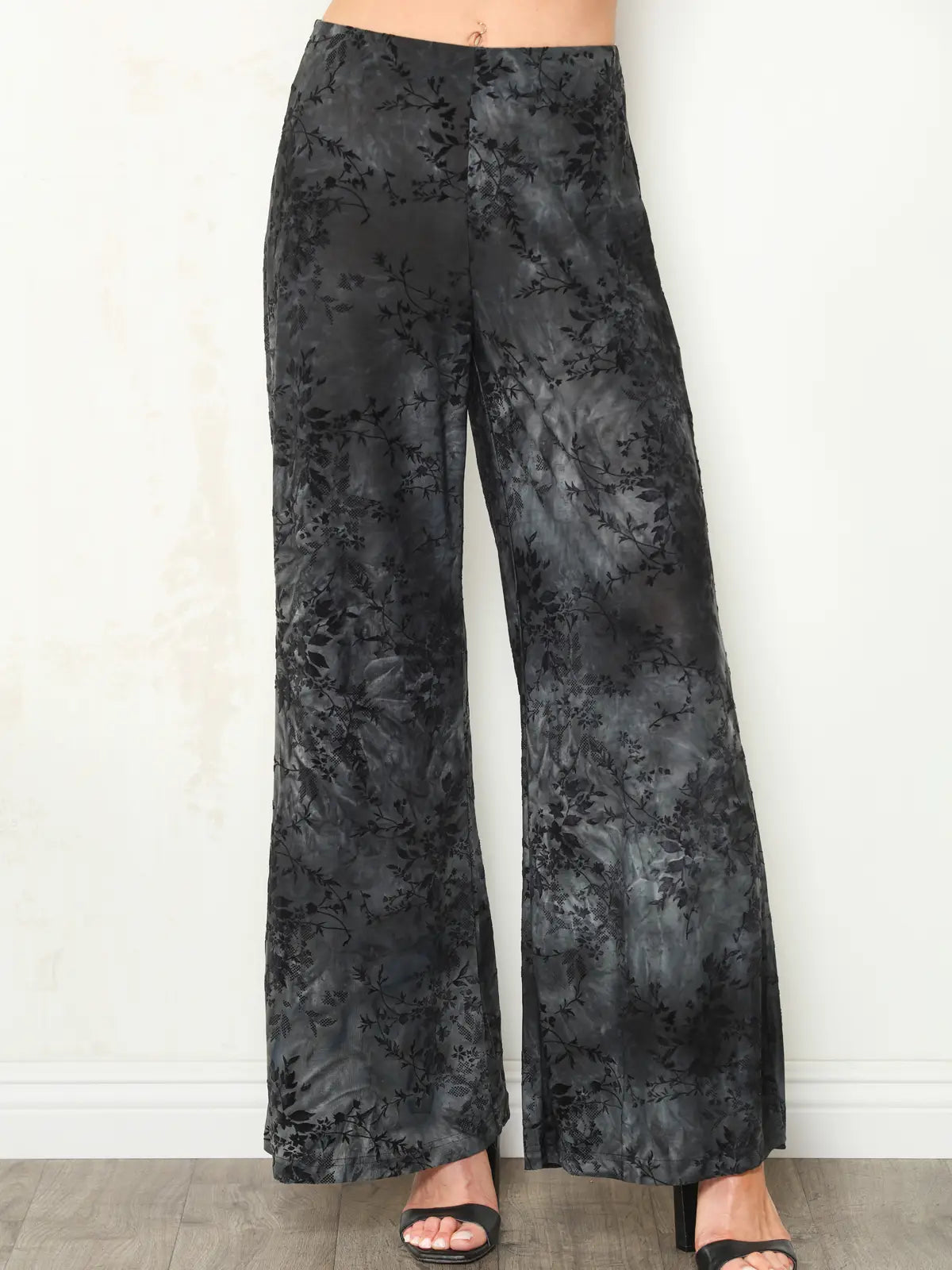 Black wide-leg pants with a subtle tonal floral jacquard pattern, shown on a model wearing black heeled sandals against a white wall.