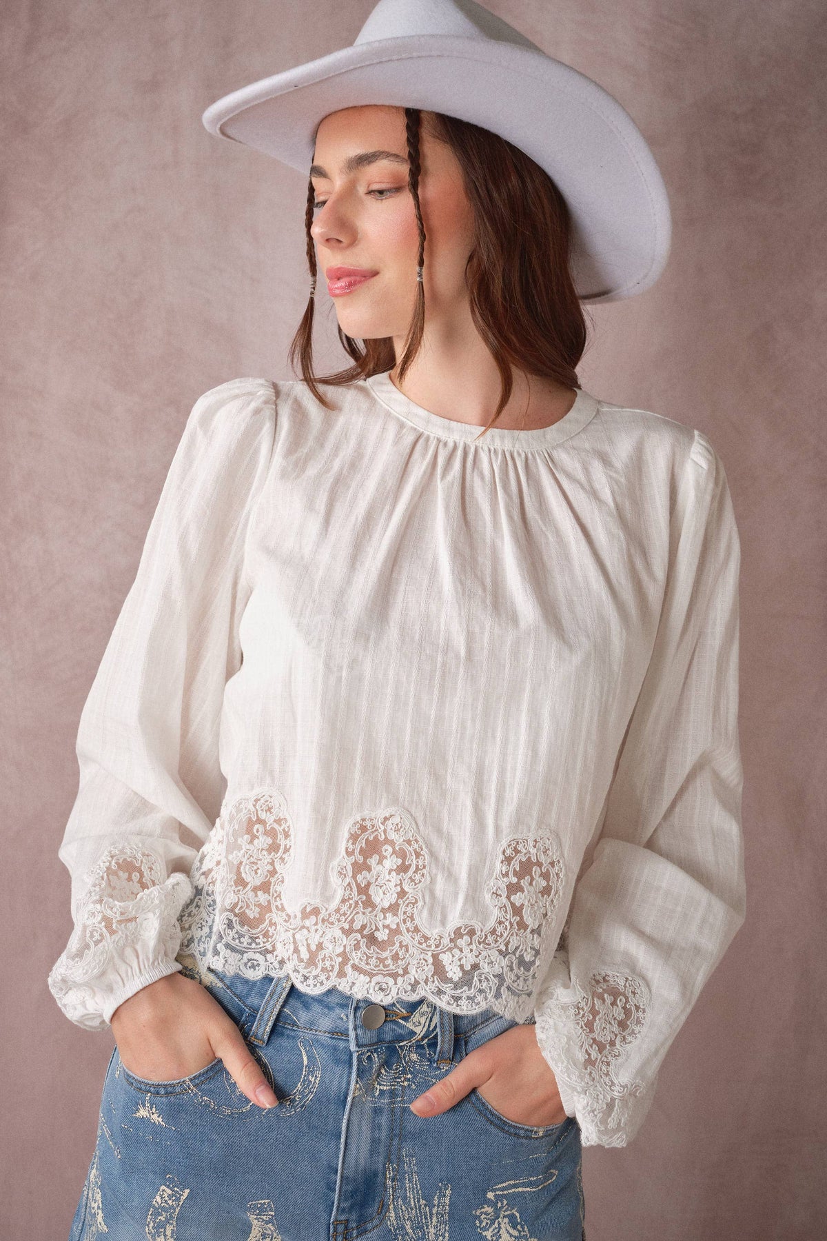 A woman wearing a cream-colored long-sleeve blouse with a gathered neckline and lace hem, paired with distressed blue jeans and a white wide-brim hat, set against a textured mauve background.