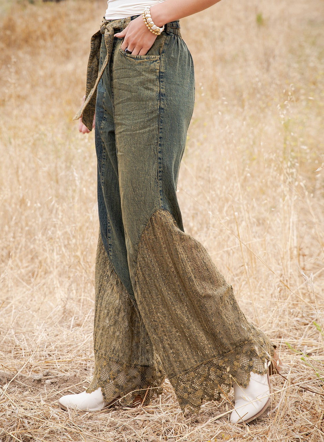 A person standing in a dry grass field wearing wide-leg patchwork jeans with a dark green denim upper section and a brown lace overlay on the lower legs, paired with white boots.