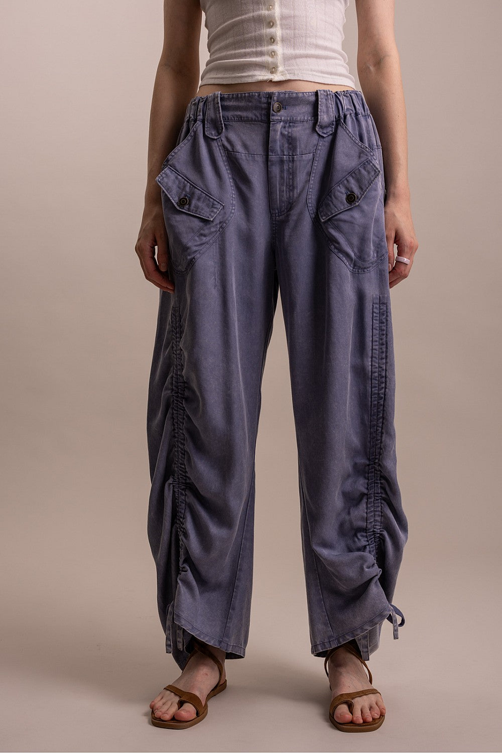 Front view of woman wearing denim blue mineral wash wide-leg cargo pants with flap pockets, drawstring ruched leg detail at hem, and elastic waist, styled with a white button-front tank and brown sandals