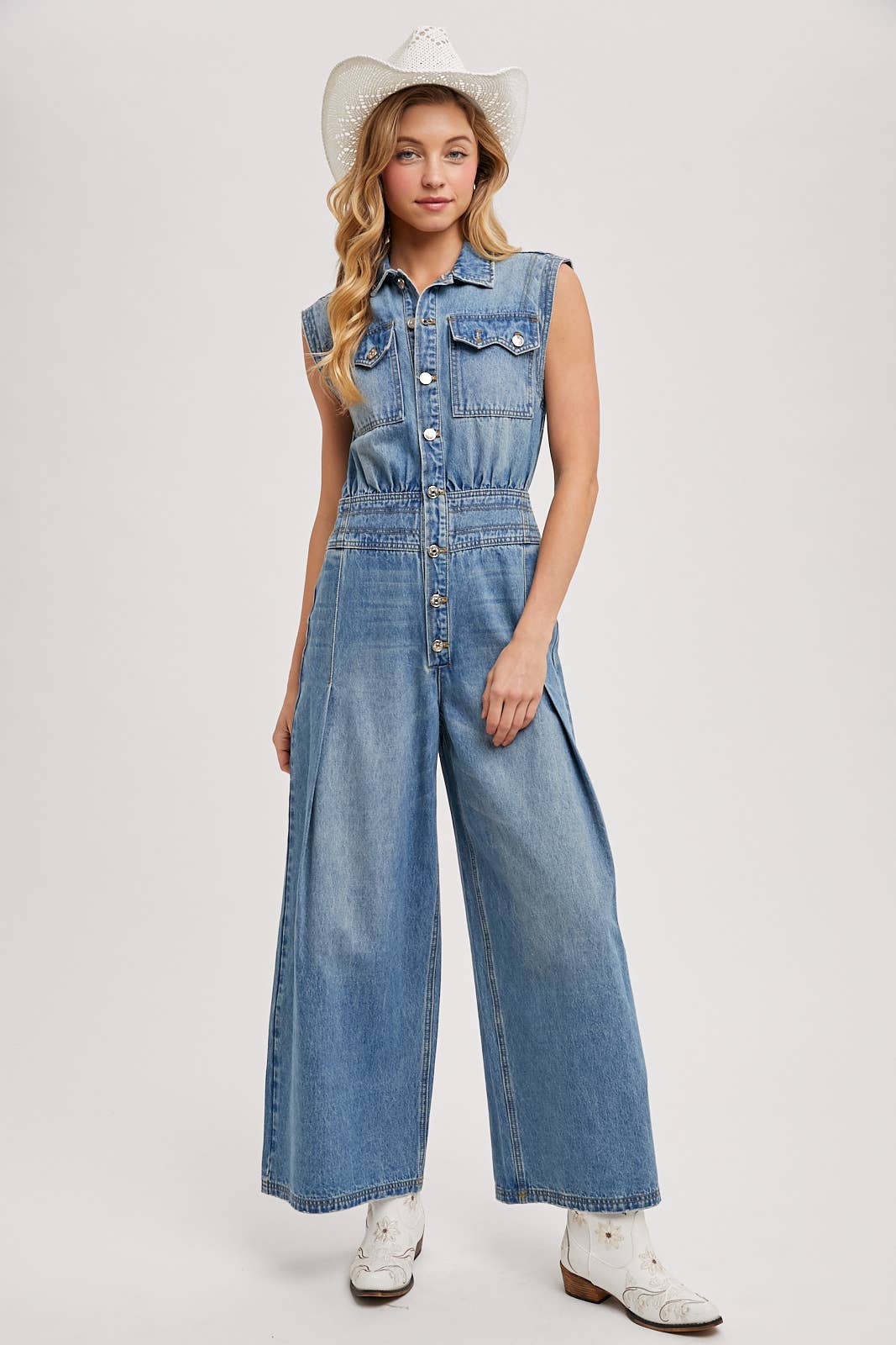 Sleeveless denim jumpsuit with collared button-front, chest flap pockets, and wide-leg palazzo silhouette, shown on model with slide sandals.