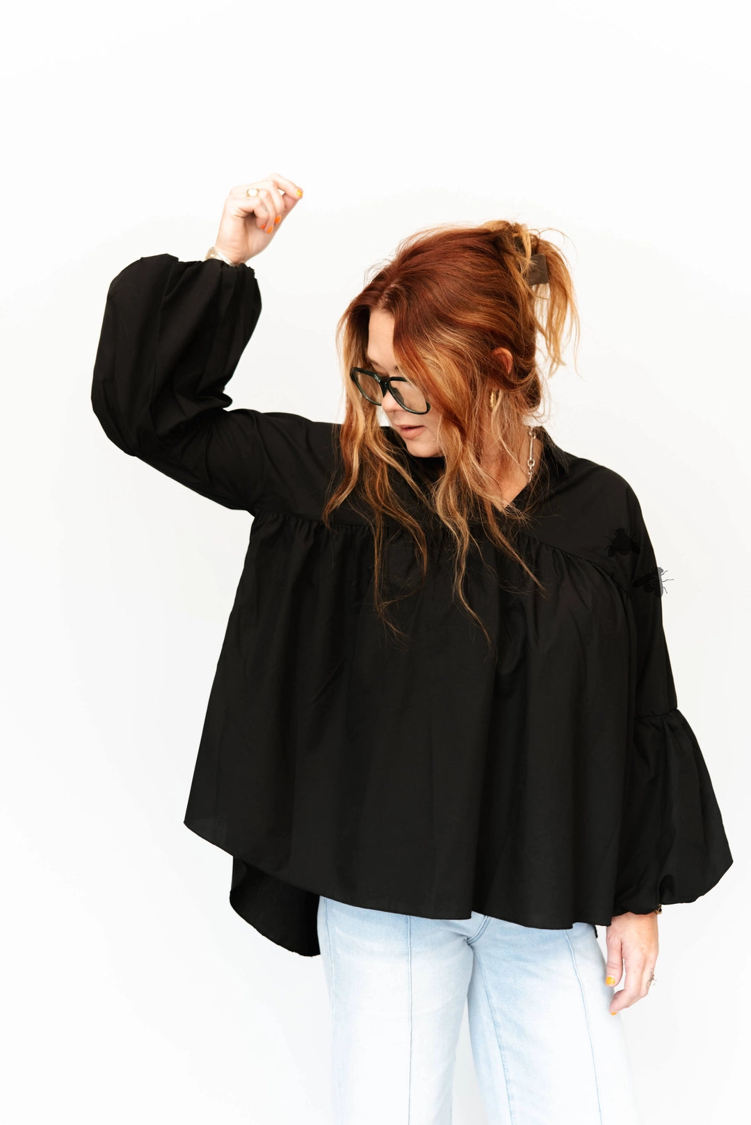 A woman with red hair and glasses wearing a loose black long-sleeve blouse with dramatic puff sleeves and light blue jeans, posing against a solid white background.