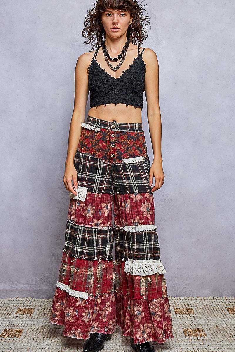 A model stands against a textured gray wall wearing a black lace crop top and highly detailed, multi-layered patchwork wide-leg pants featuring floral, plaid, and lace patterns in red, black, and beige tones.