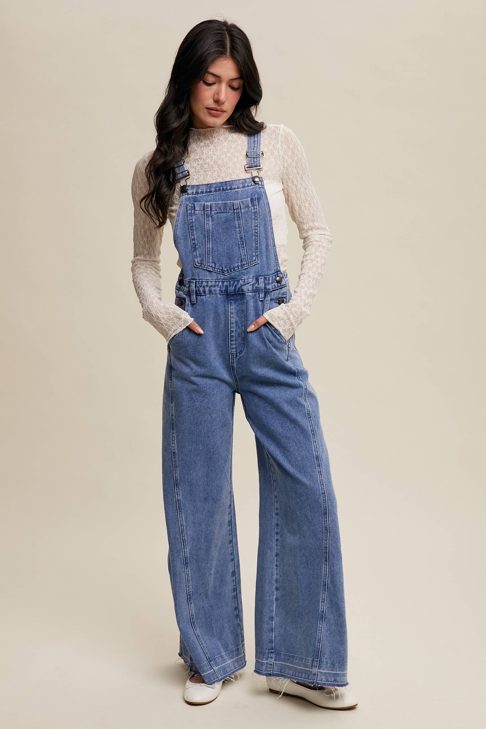 Light-wash wide-leg denim overalls with adjustable straps, front bib pocket, side buttons, and slant pockets, finished with a raw hem and layered over a cream long-sleeve top.
