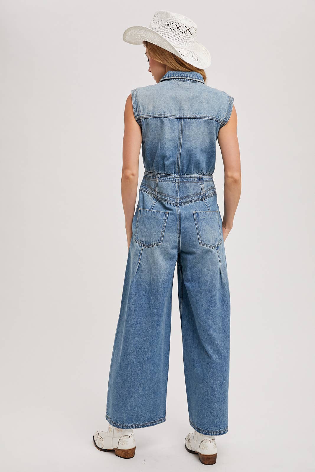 Back view of sleeveless denim jumpsuit with collared neckline, defined waist, back patch pockets, and wide-leg pants, worn with brown slide sandals.