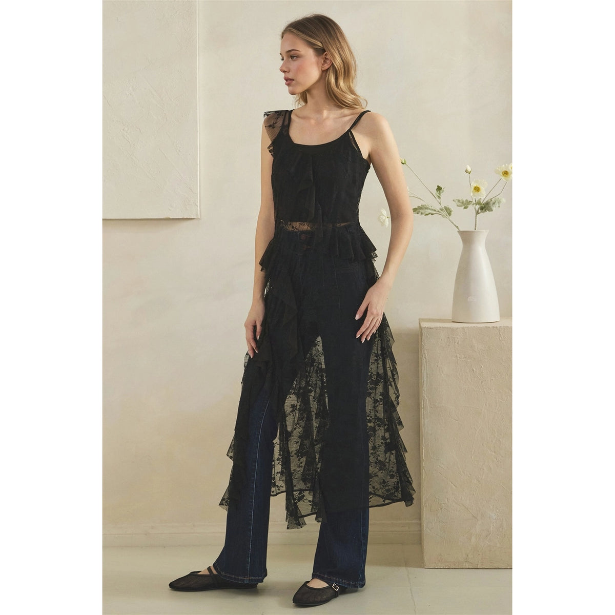 A woman models a black sheer lace ruffled duster over dark wide-leg jeans, standing in a minimalist beige room with a white vase on a pedestal.