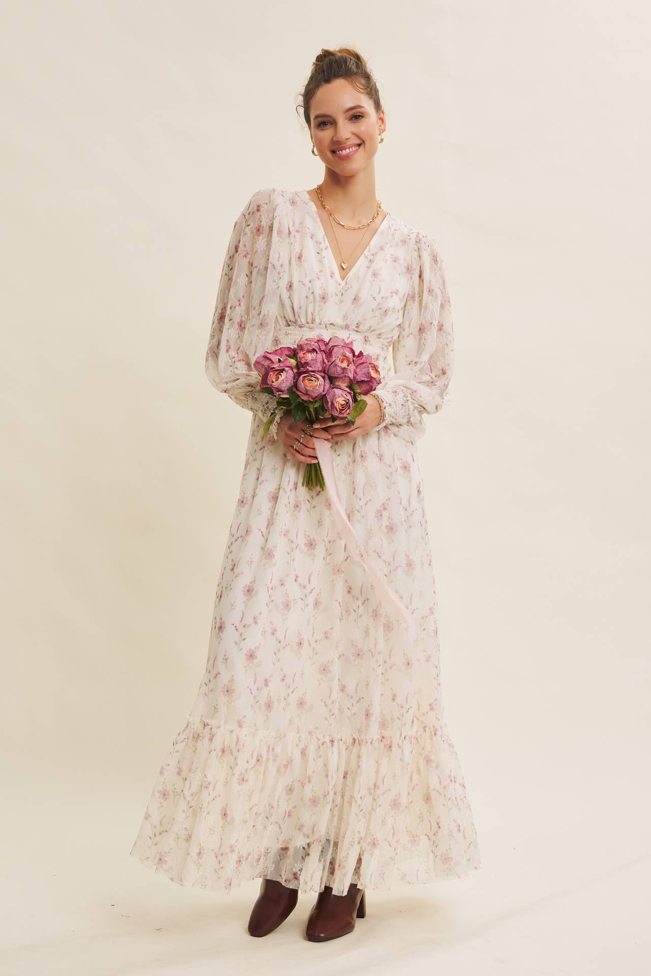 Front view — cream floral chiffon maxi dress with V-neck, sheer long sleeves, smocked waist, ruffle hem; model holding pink bouquet.