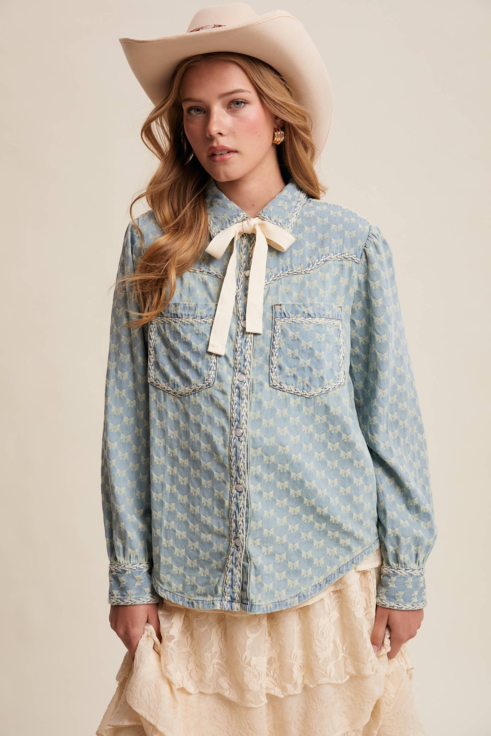 Light-wash denim blouse with cream bow-tie collar, tonal mini-bow pattern, braided contrast stitching, snap front, and two chest pockets.