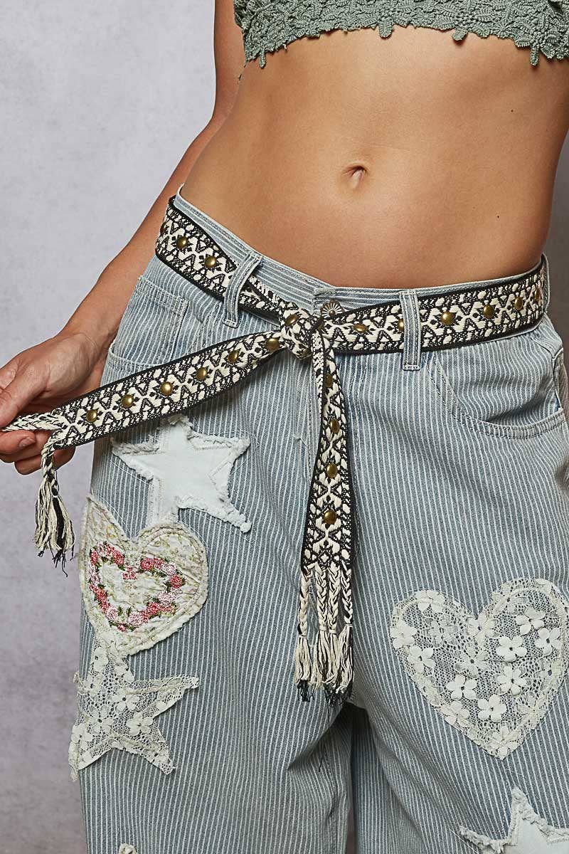 Close-up of a woven black and beige boho belt with stud accents and tassel ends, styled around striped denim jeans with lace patches.