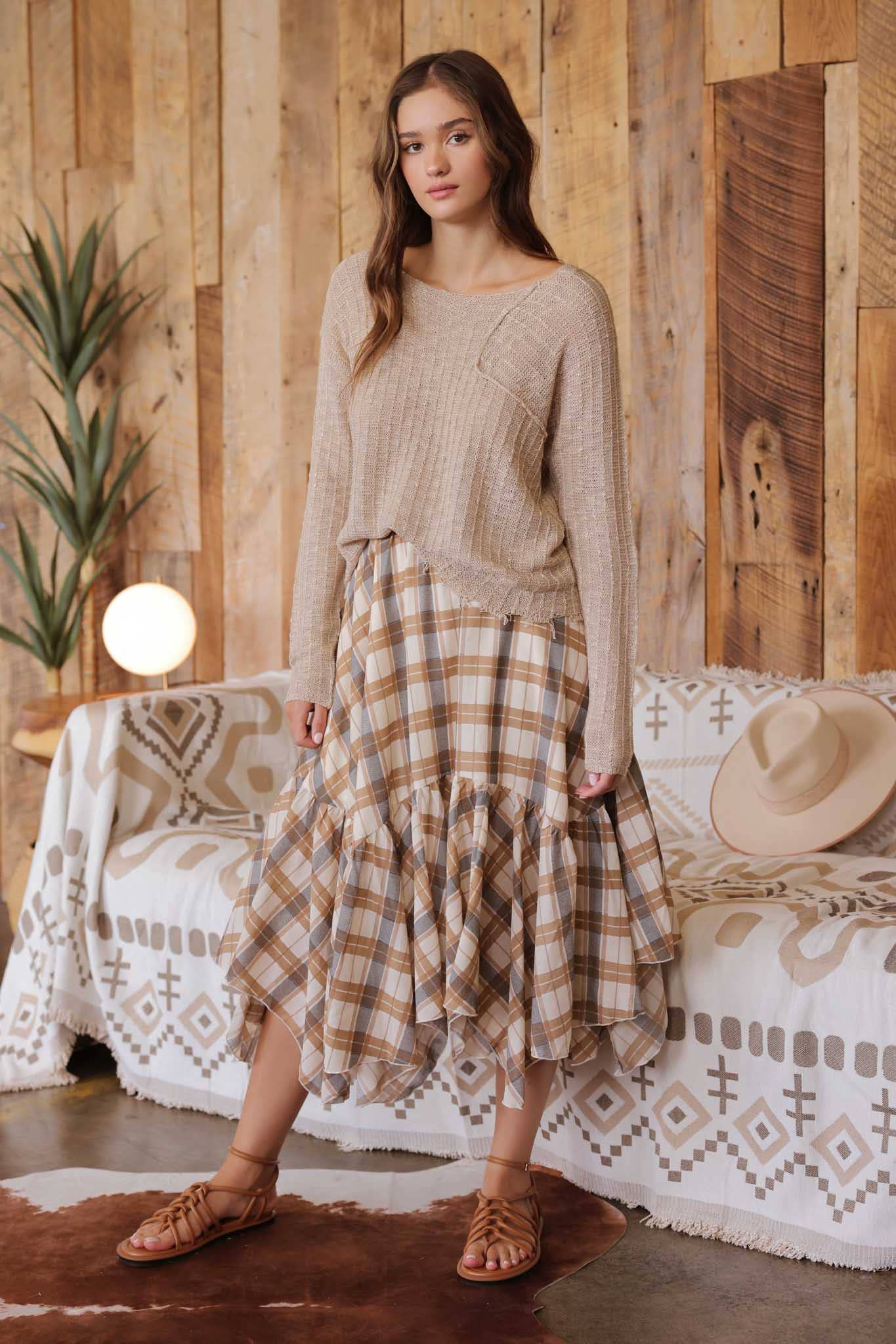 Woman wearing a beige sweater and plaid skirt standing in a rustic room with wooden walls and a couch.