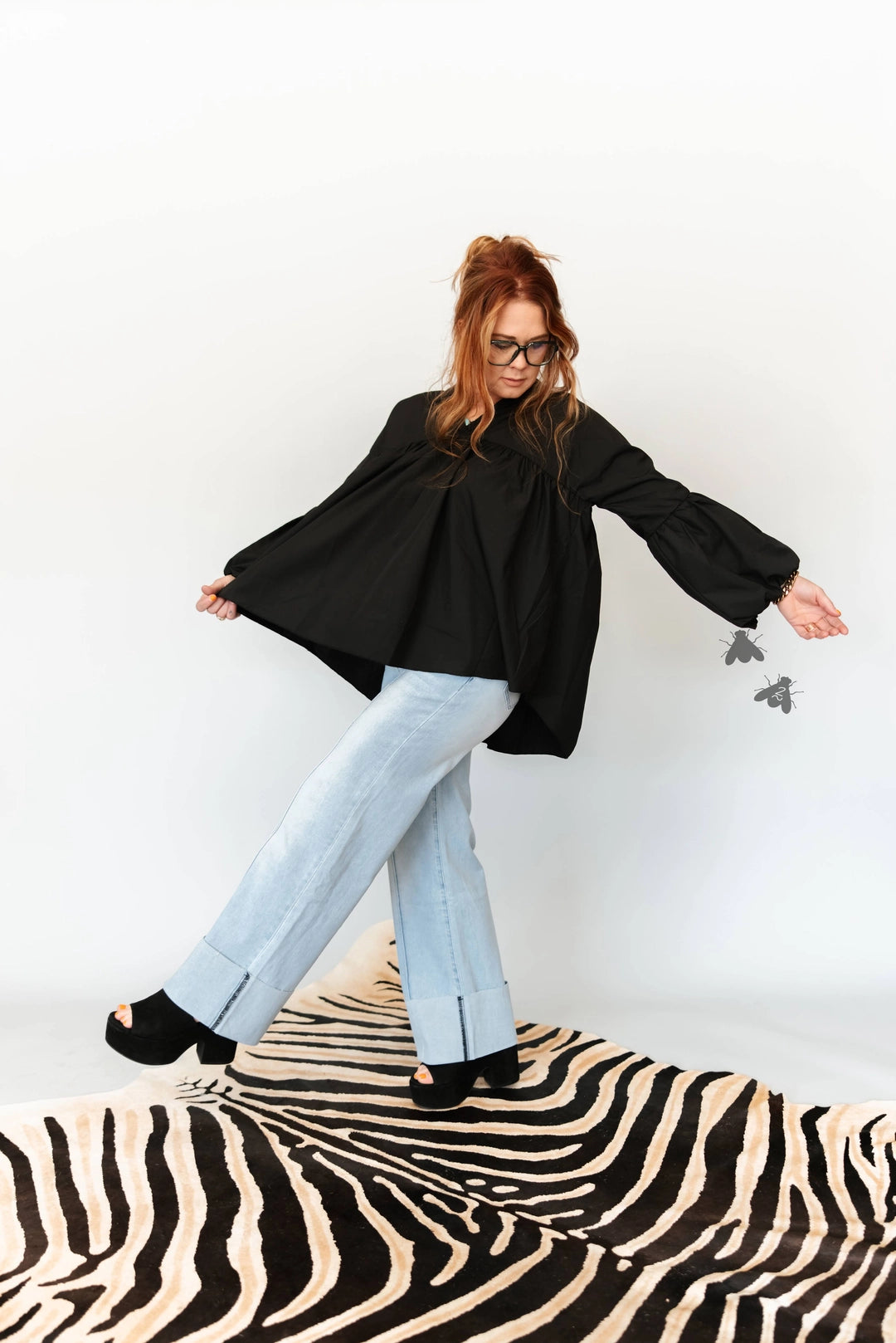A woman wearing a black oversized long-sleeve blouse, light-wash wide-leg jeans, and black platform sandals poses on a black and beige zebra-print rug against a white studio background.