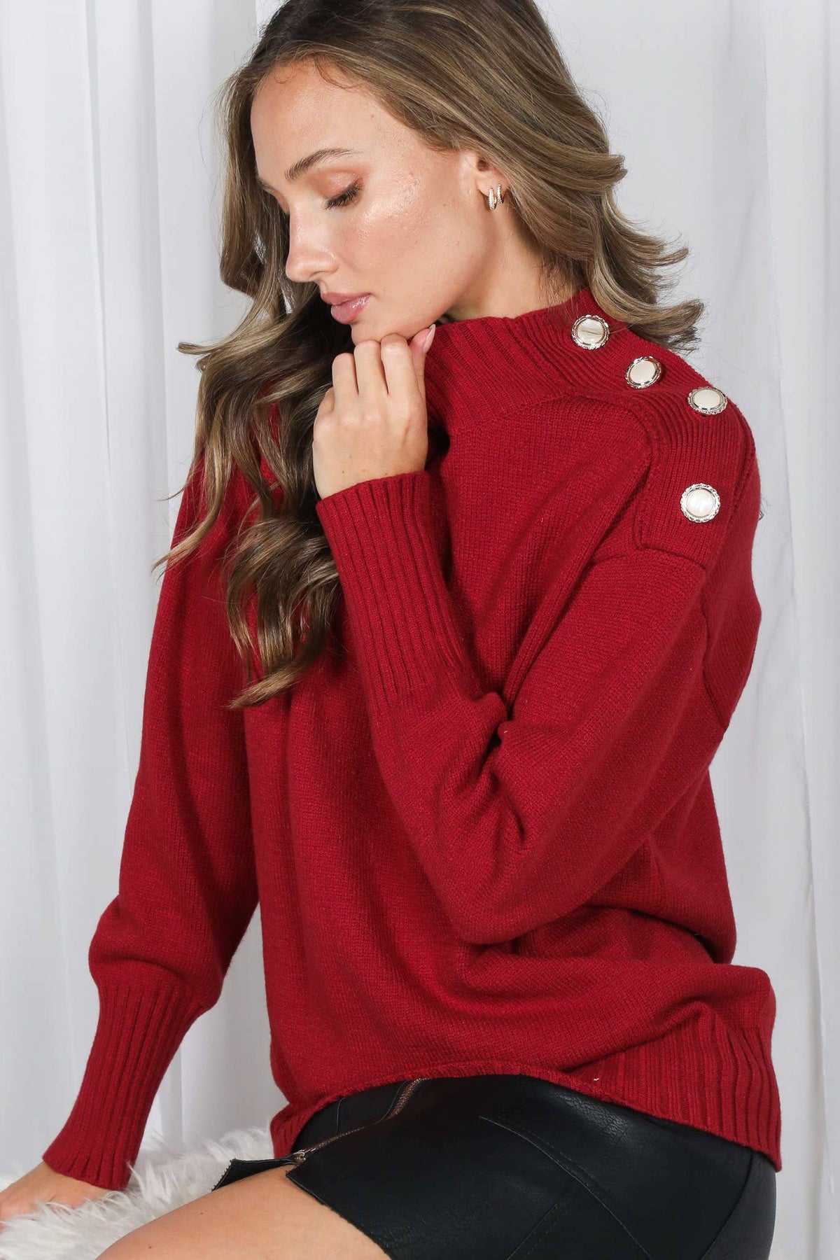 A woman wearing a red knit sweater with decorative silver-toned buttons on the shoulders, paired with black shorts, posed against a white curtain background.