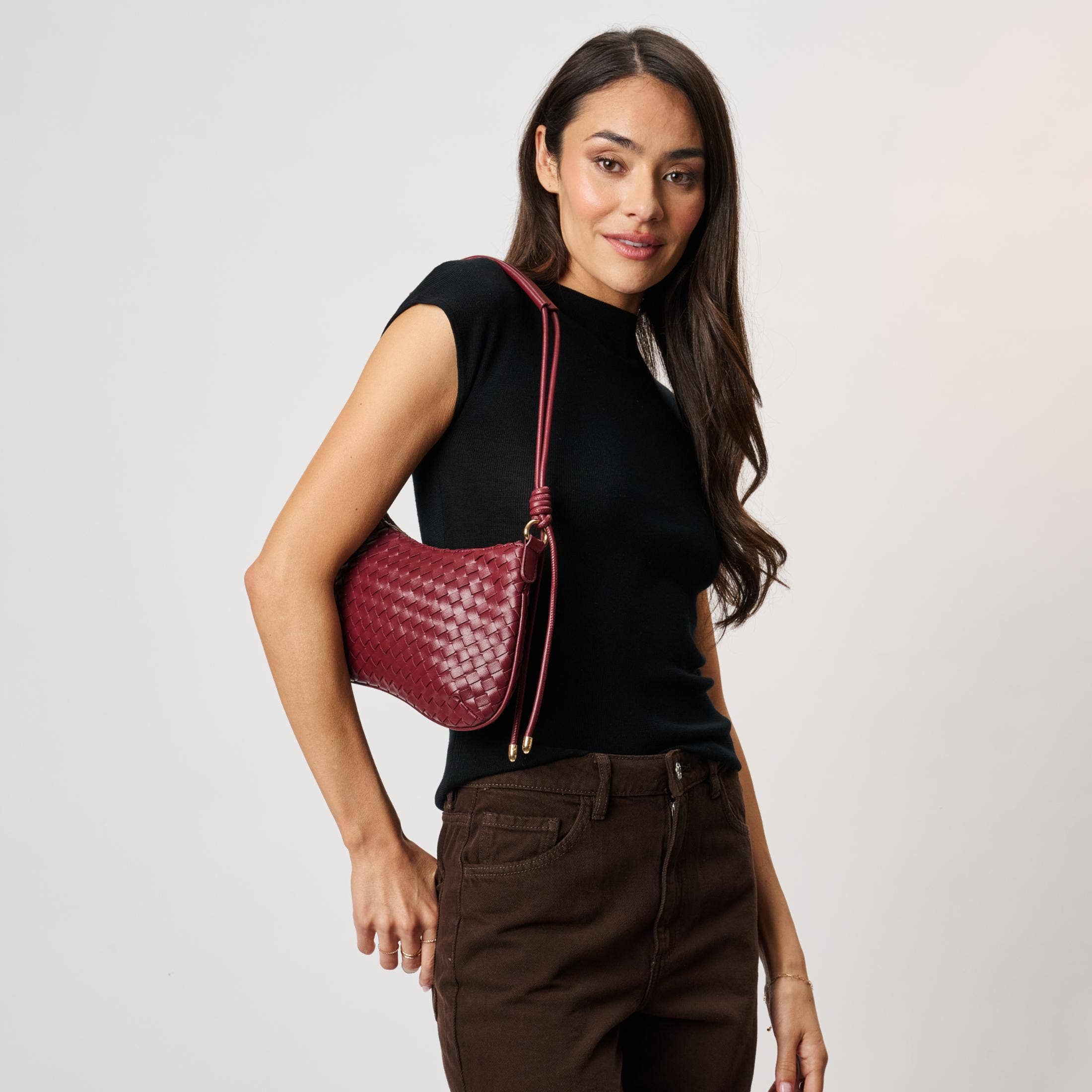 Cindy Woven Crescent Shoulder Bag in Burgundy by Moda Luxe — model shoulder carry view showing woven vegan leather crescent bag with knotted strap, gold-tone hardware and zip closure, styled with black top and dark brown trousers