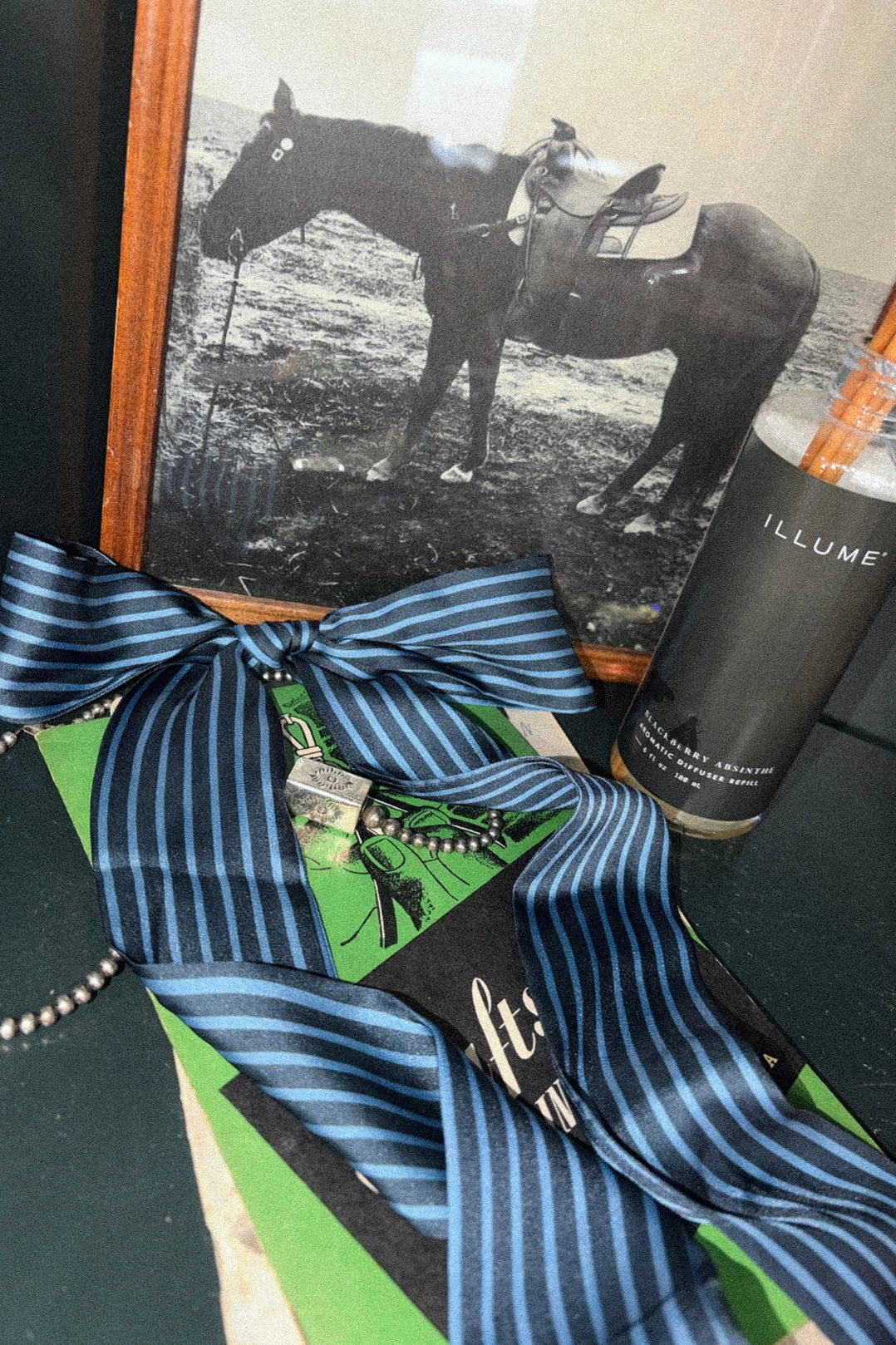 A blue and navy striped necktie with a matching striped bow and ribbon, displayed on top of a green book next to a black bottle labeled ILLUME. A framed black-and-white photo of a horse rider is in the background.