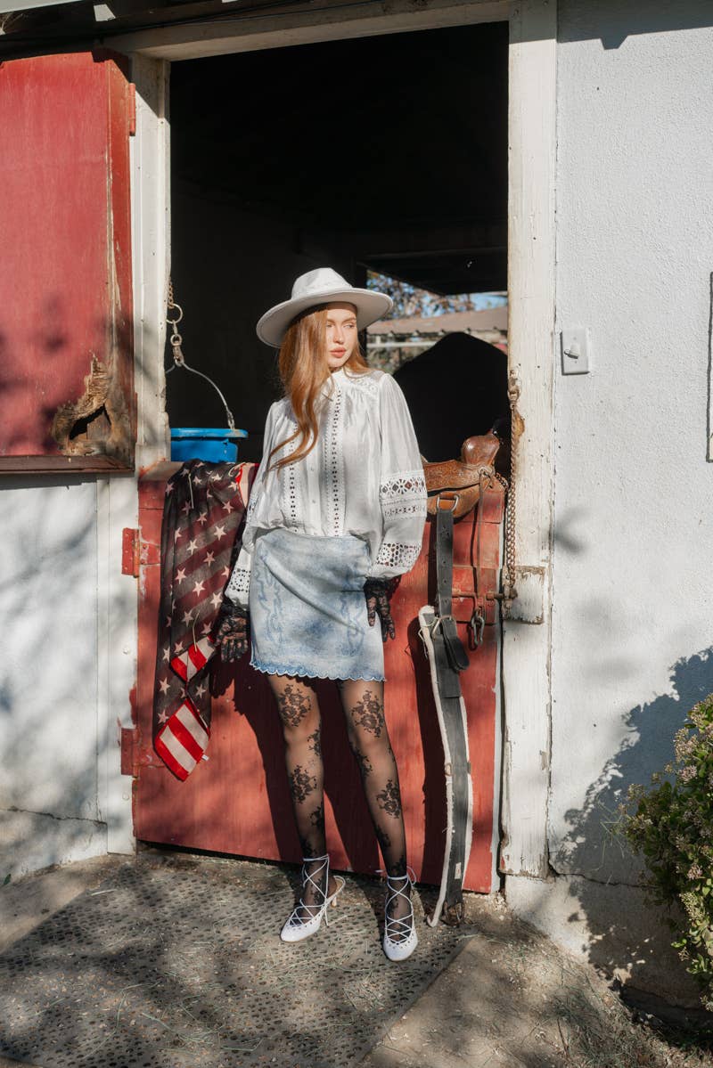 Woman standing in front of a red barn door with a cowboy hat and lace top.