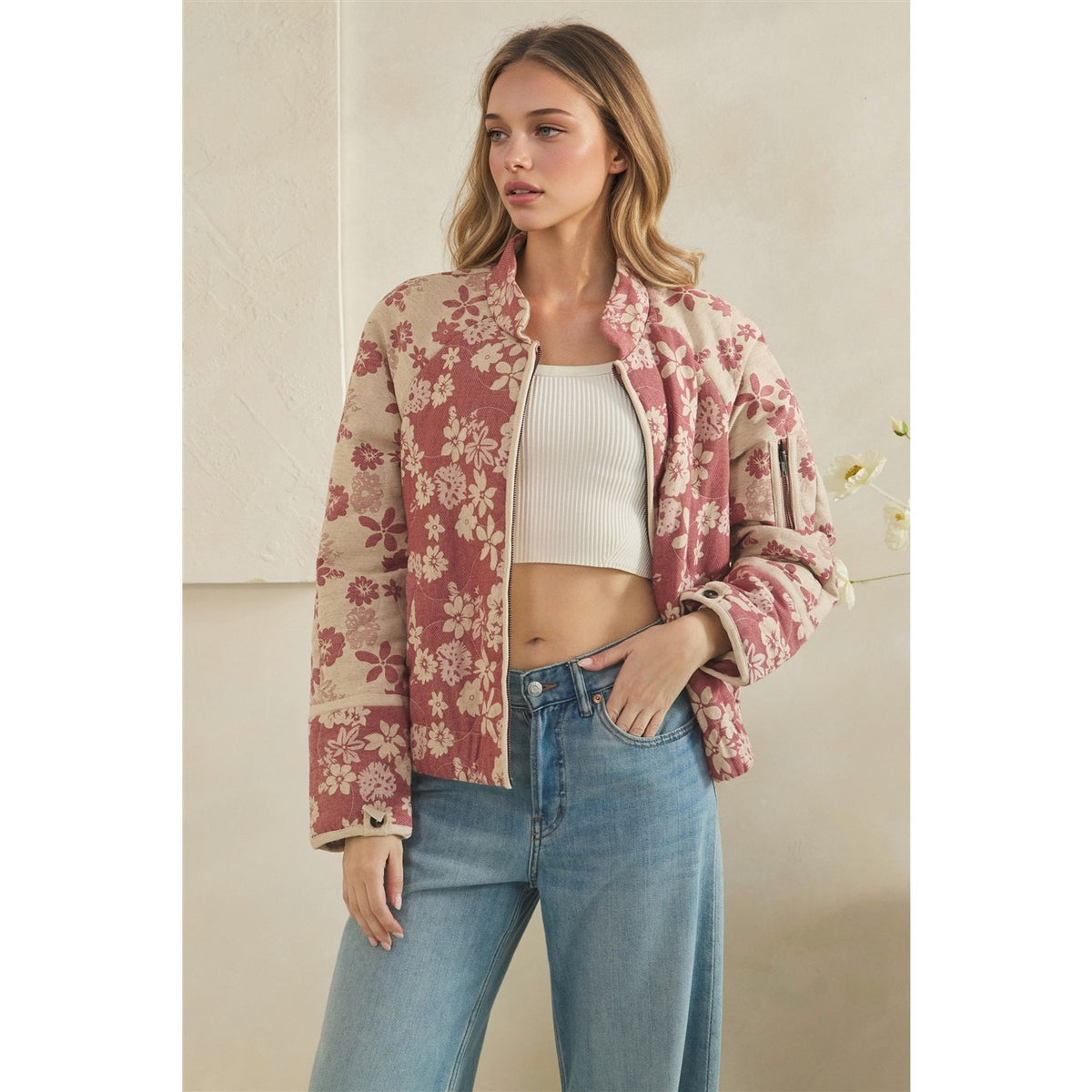 A woman models a cropped floral bomber jacket with a pink and beige pattern, worn over a white ribbed crop top and blue jeans against a neutral beige wall.