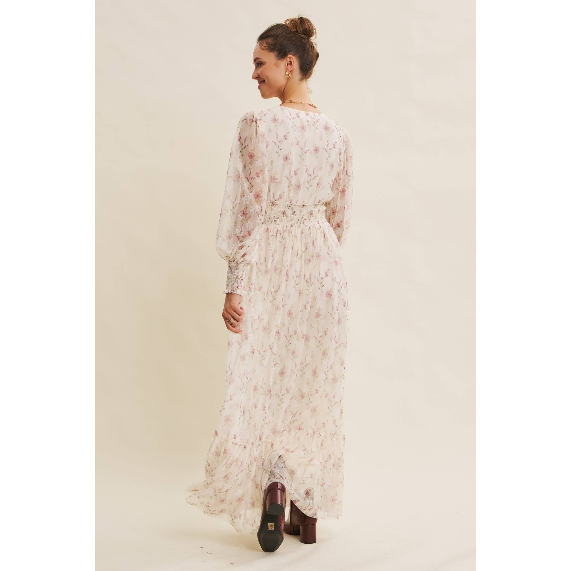 Back view — cream floral chiffon maxi dress with V-neck, sheer long sleeves, smocked waist, and ruffle hem; styled with brown boots.