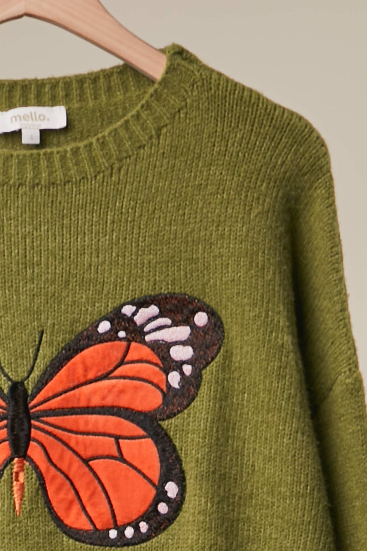 Olive green knit sweater with a large embroidered monarch butterfly on the front, hanging on a wooden hanger against a neutral beige background.