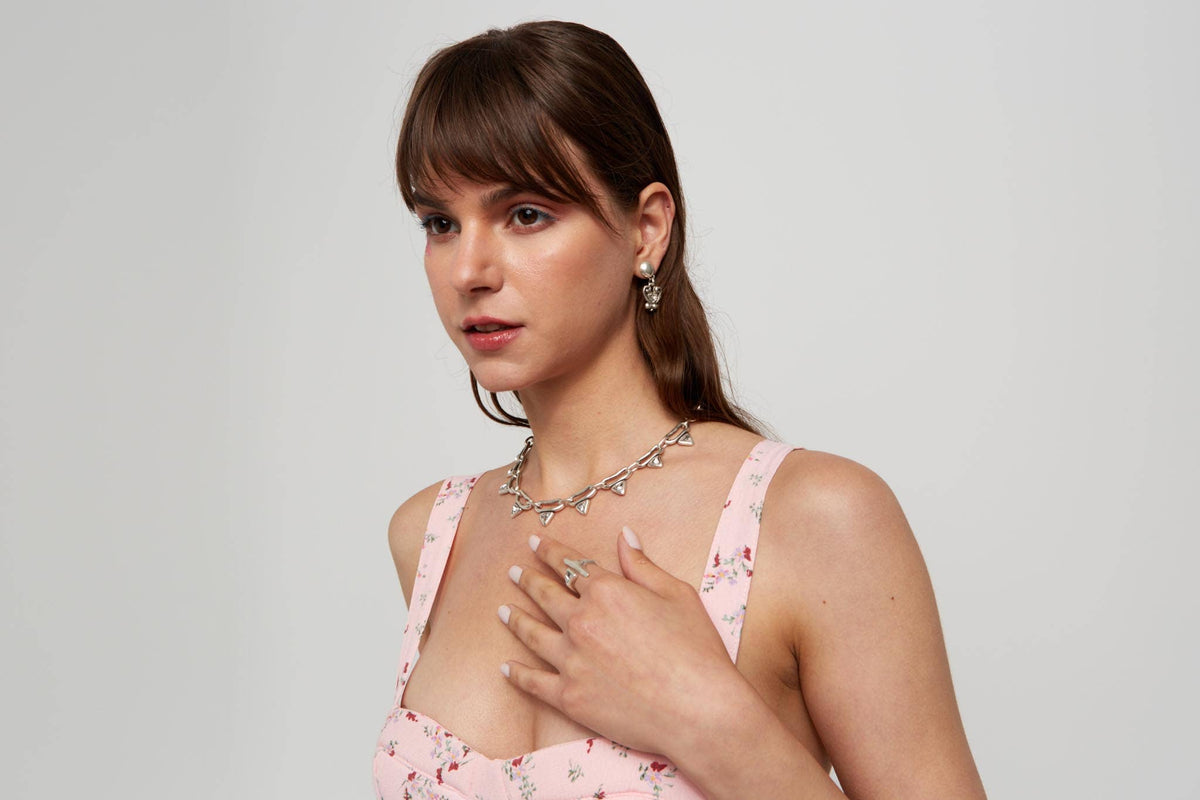 A woman with brown hair and bangs models a silver-toned leaf choker necklace and matching leaf earrings, wearing a light pink floral spaghetti-strap dress against a light gray background.