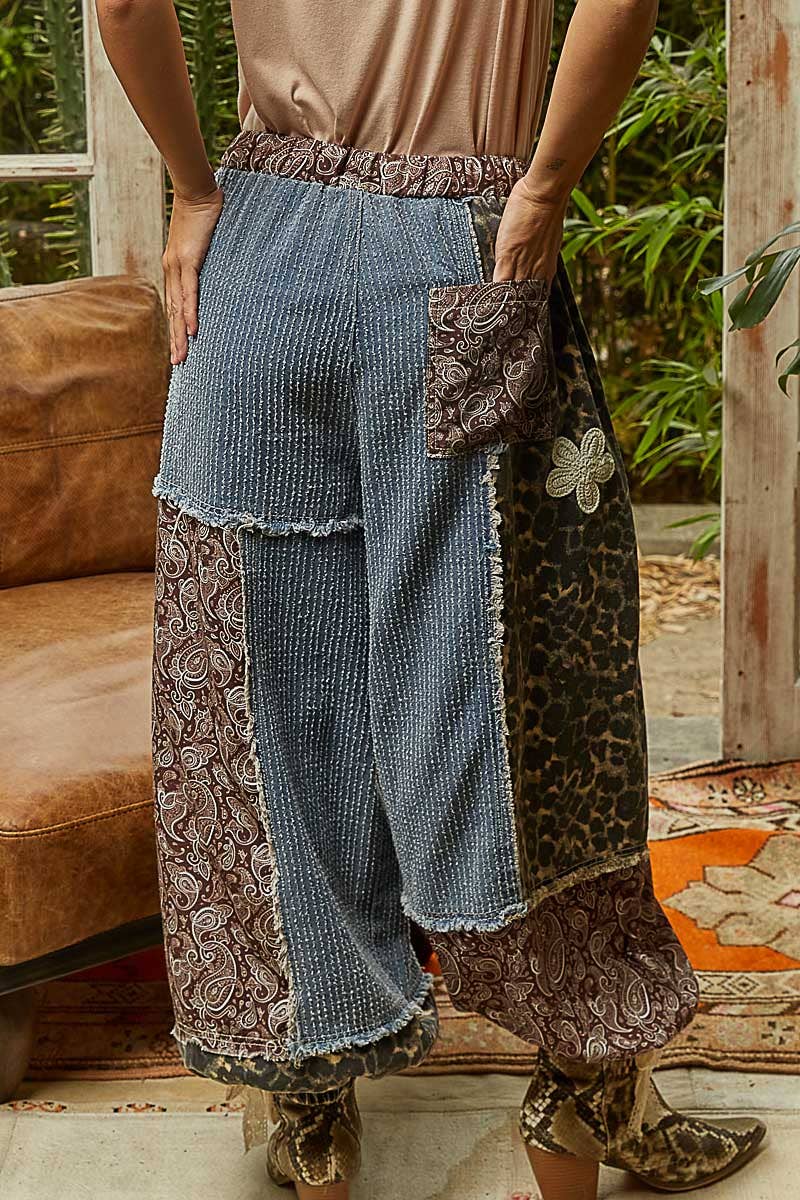 Person wearing a unique patterned outfit with wide-leg pants and a brown top, standing in a room with a couch and plants.