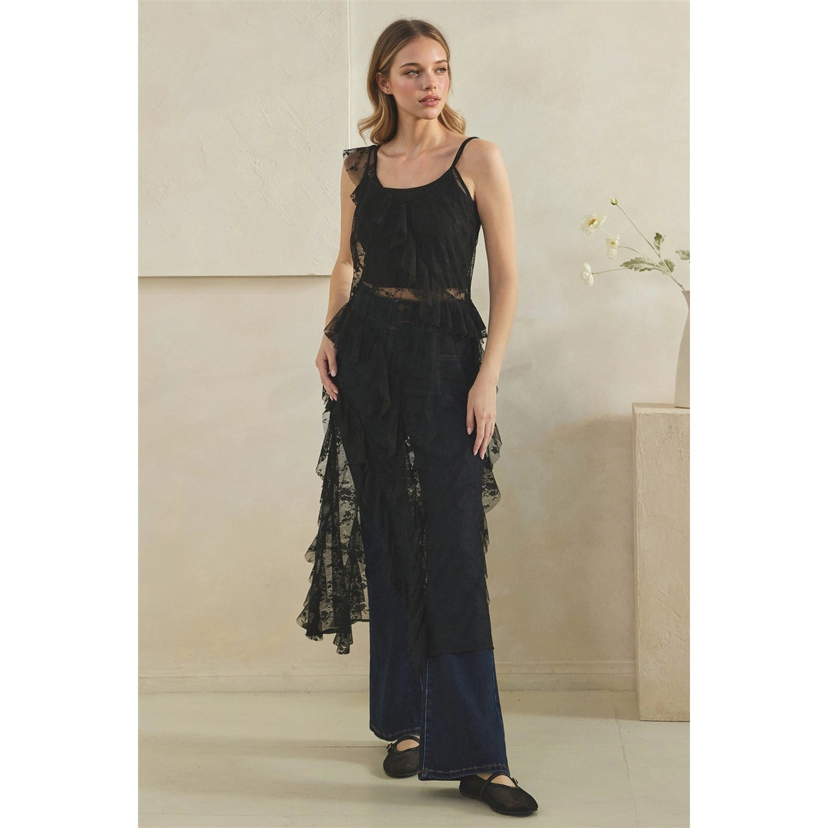 A woman models a black lace sleeveless duster with dark wide-leg jeans in a minimalist beige interior setting.