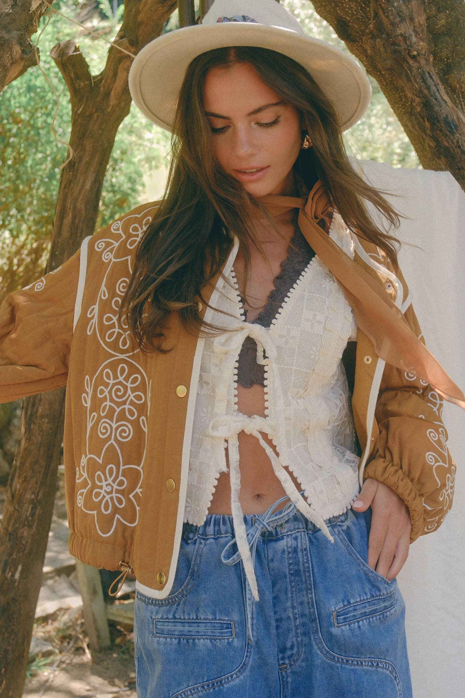 Boho caramel-brown jacket with cream floral swirl embroidery, ivory piping, and snap buttons, styled over a lace tie-front top with a scarf and felt hat outdoors.