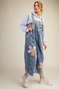 Three-quarter view of woman wearing light blue corduroy wide-leg overalls with large floral patchwork detail, paired with a white hoodie and white sneakers