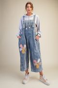 Front view of woman wearing light blue washed corduroy wide-leg overalls with floral patchwork appliqués, styled with a white hoodie and white sneakers
