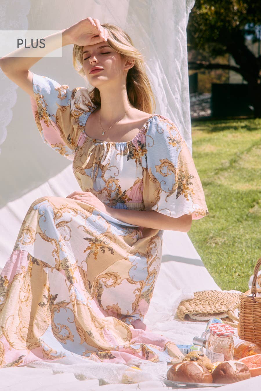 Woman in a floral dress sitting outdoors with a picnic basket and food items.