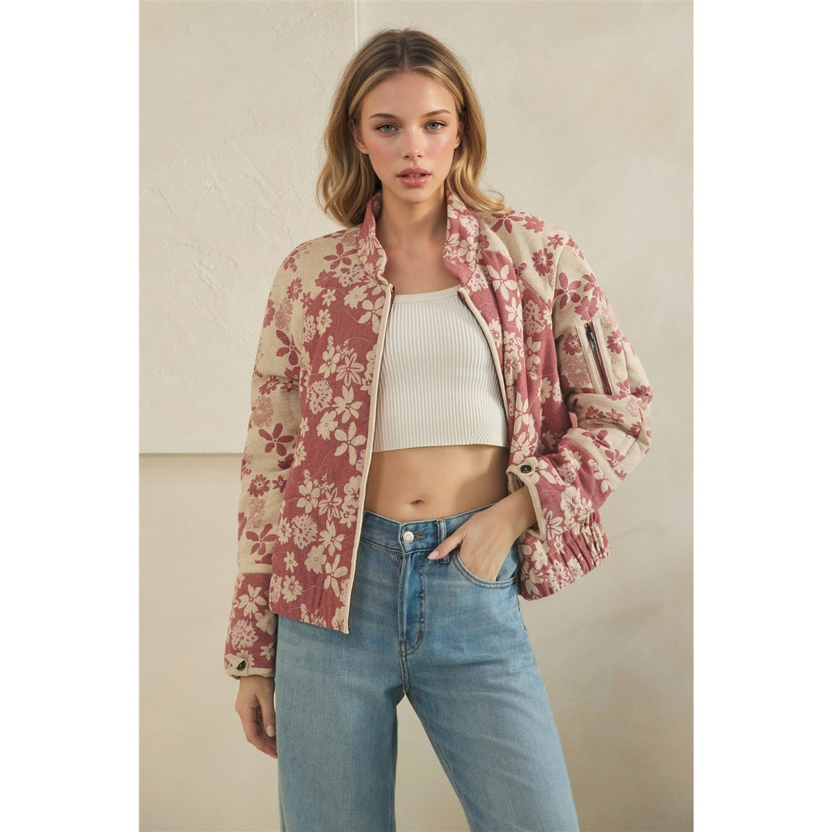 A woman models a cropped, lightweight bomber-style jacket with a dusty rose and beige floral pattern, worn open over a white ribbed crop top and blue jeans against a neutral beige wall.