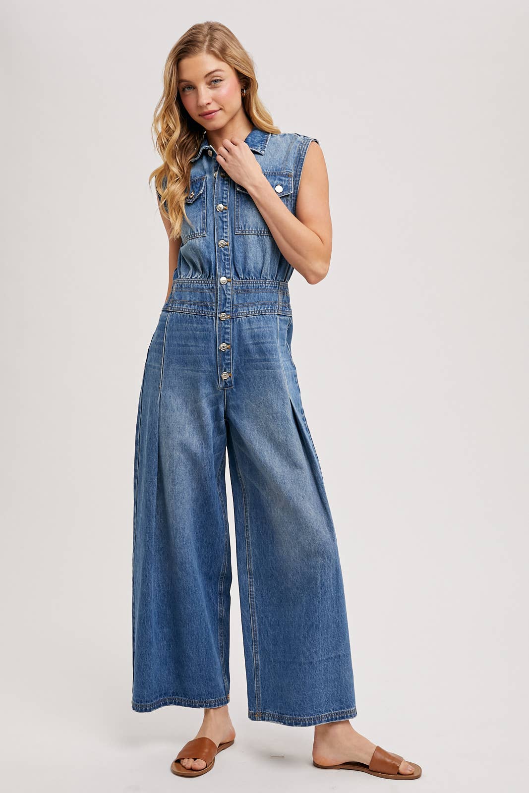 Sleeveless denim jumpsuit with collared button-front, chest flap pockets, and wide-leg palazzo silhouette, shown on model with slide sandals.