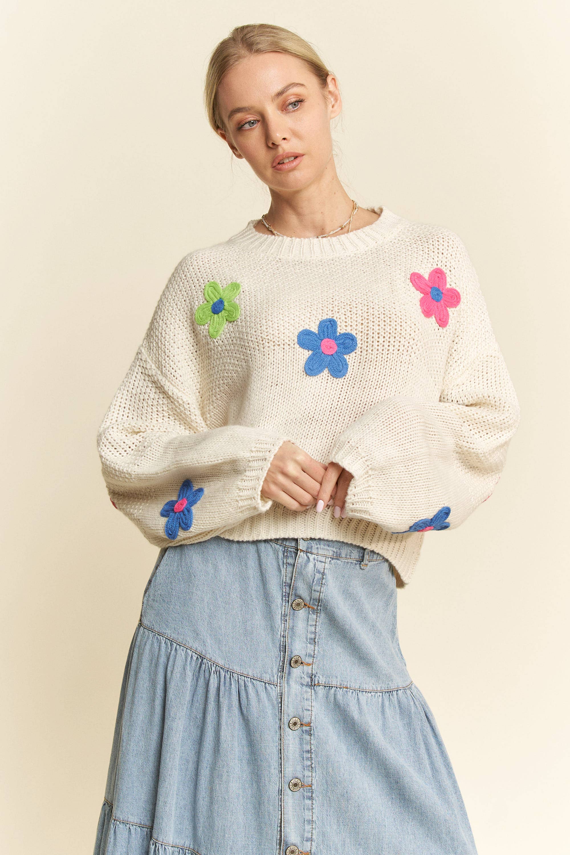 Cream knit sweater with colorful 3D floral appliqués in pink, blue, and green, styled with a button-front light denim skirt.