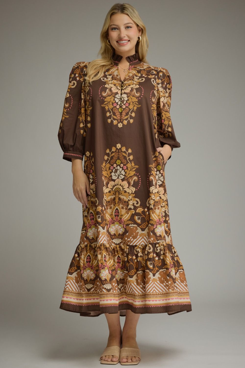 A woman modeling a brown midi dress with a detailed gold, beige, and white floral and geometric pattern, set against a solid gray studio background.