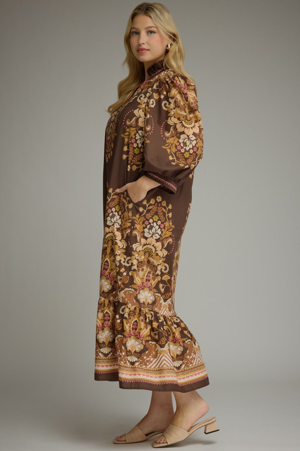 A woman modeling a brown long-sleeve maxi dress with an intricate floral and paisley print in beige, gold, and rust tones, paired with beige heeled sandals against a solid gray studio background.
