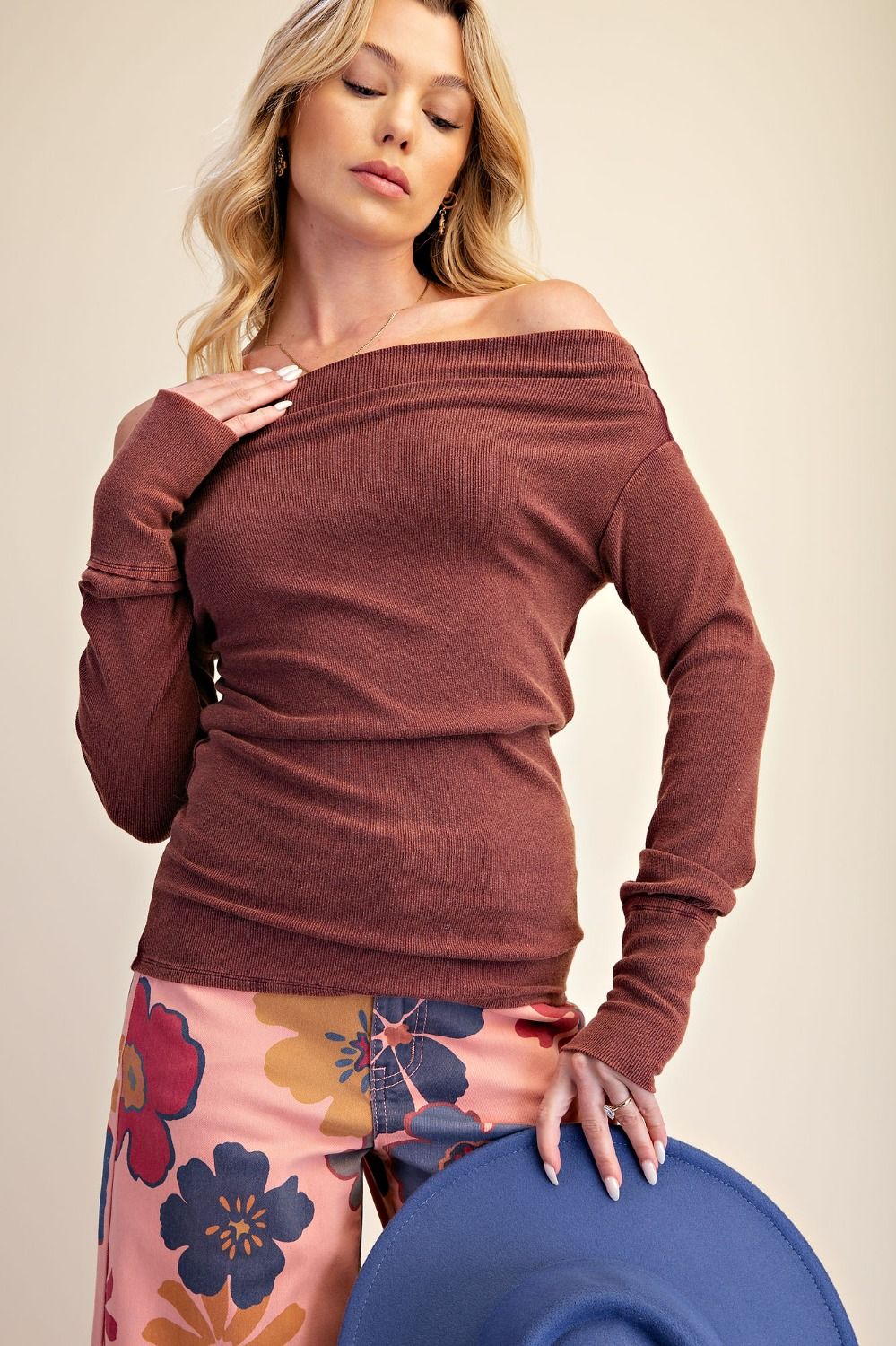 A woman wearing a rust-brown off-the-shoulder long-sleeve knit top and pink floral pants, holding a blue hat against a plain beige studio background.