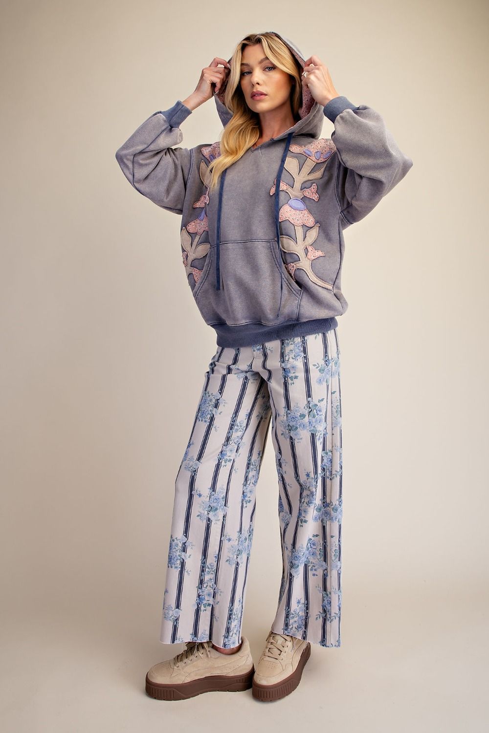 Full-length front view of woman in slate blue floral patch mineral wash hoodie with balloon sleeves and drawstring hood, styled with floral stripe wide-leg pants and platform sneakers
