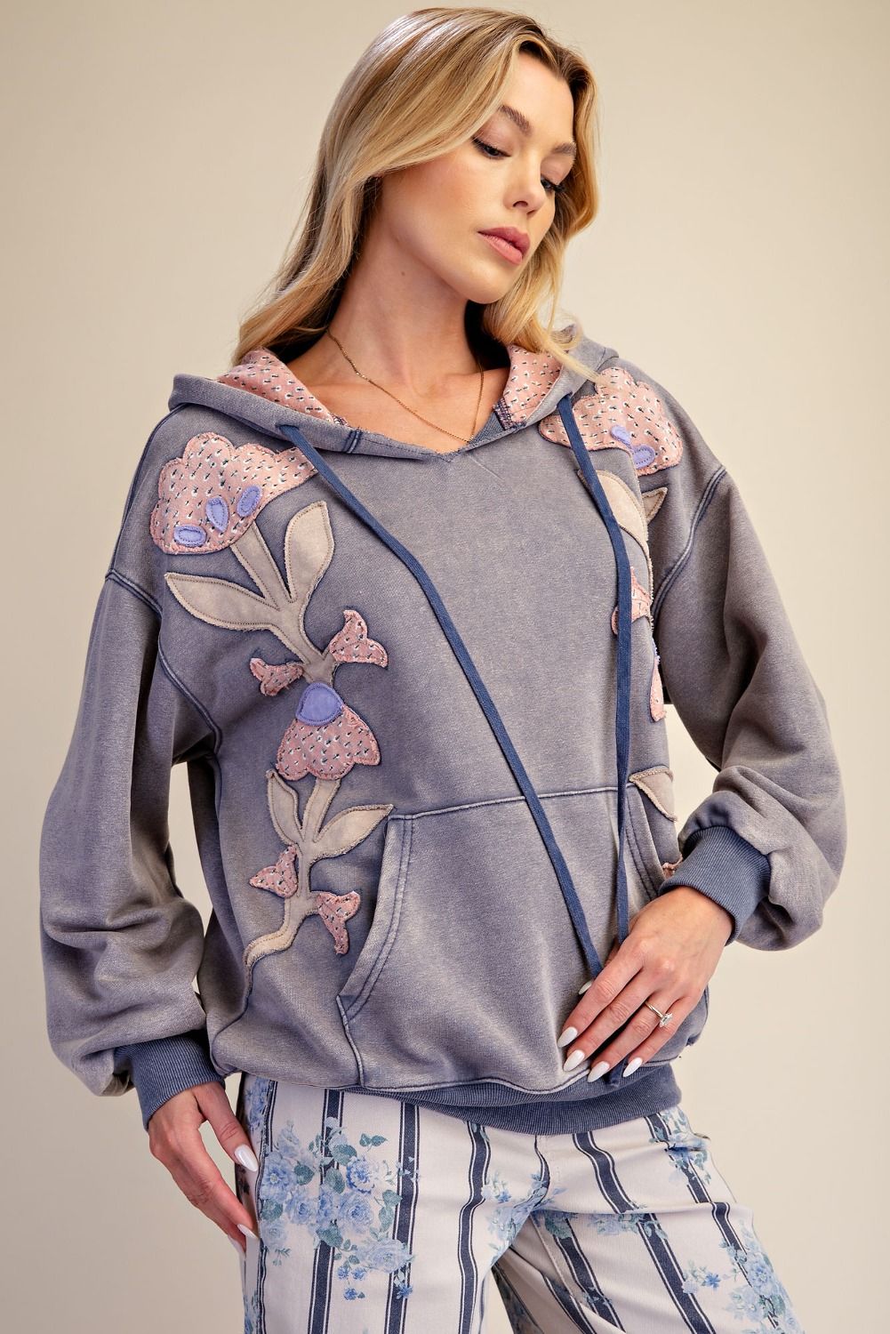 Close-up front view of woman wearing slate blue mineral wash hoodie with large floral patchwork appliqués and contrast-printed hood lining, paired with floral stripe wide-leg pants