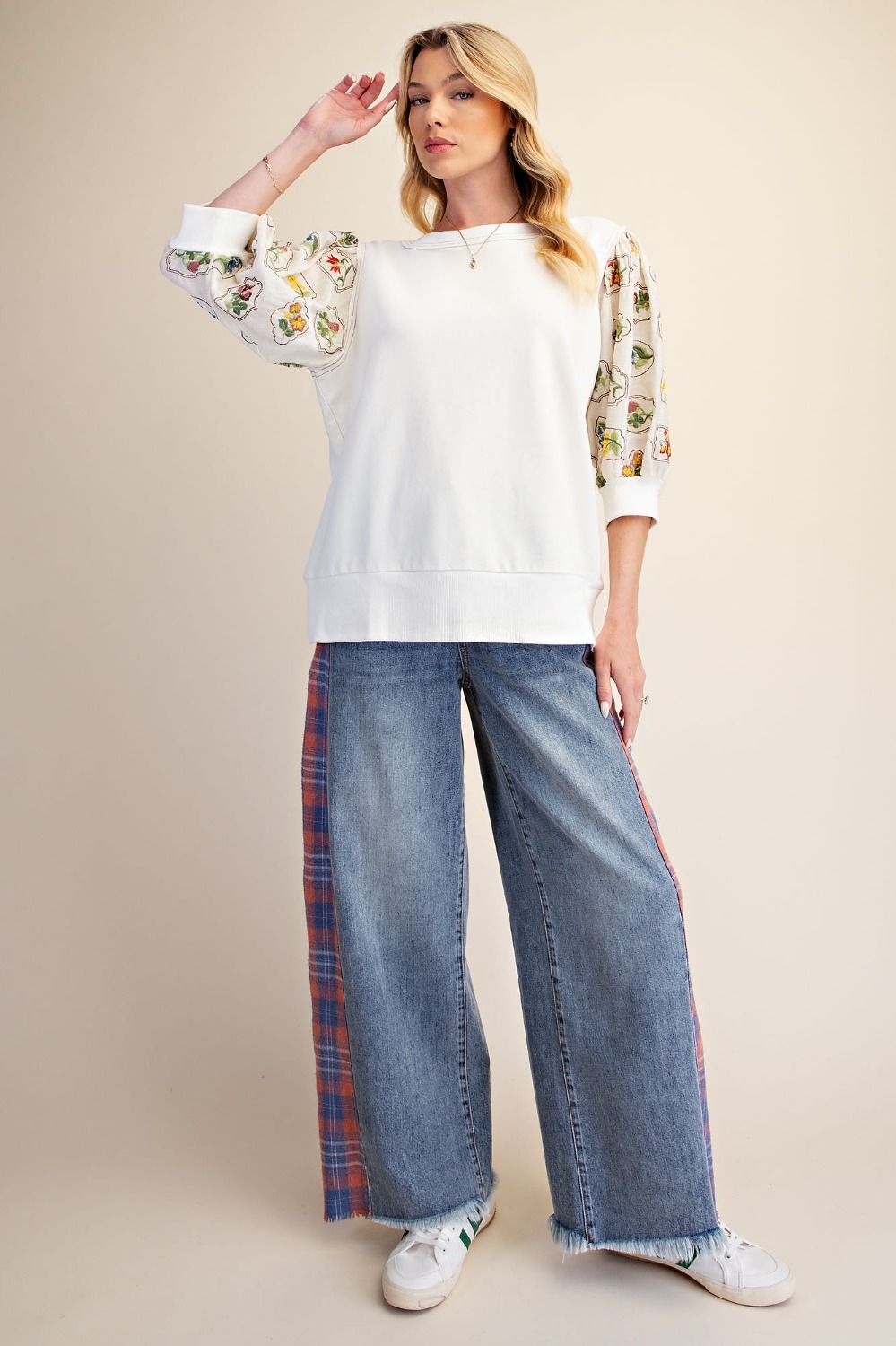 A woman modeling a white crewneck sweatshirt with floral embroidered sleeves, paired with wide-leg blue jeans featuring red and blue plaid side panels, and white sneakers, against a solid beige studio background.