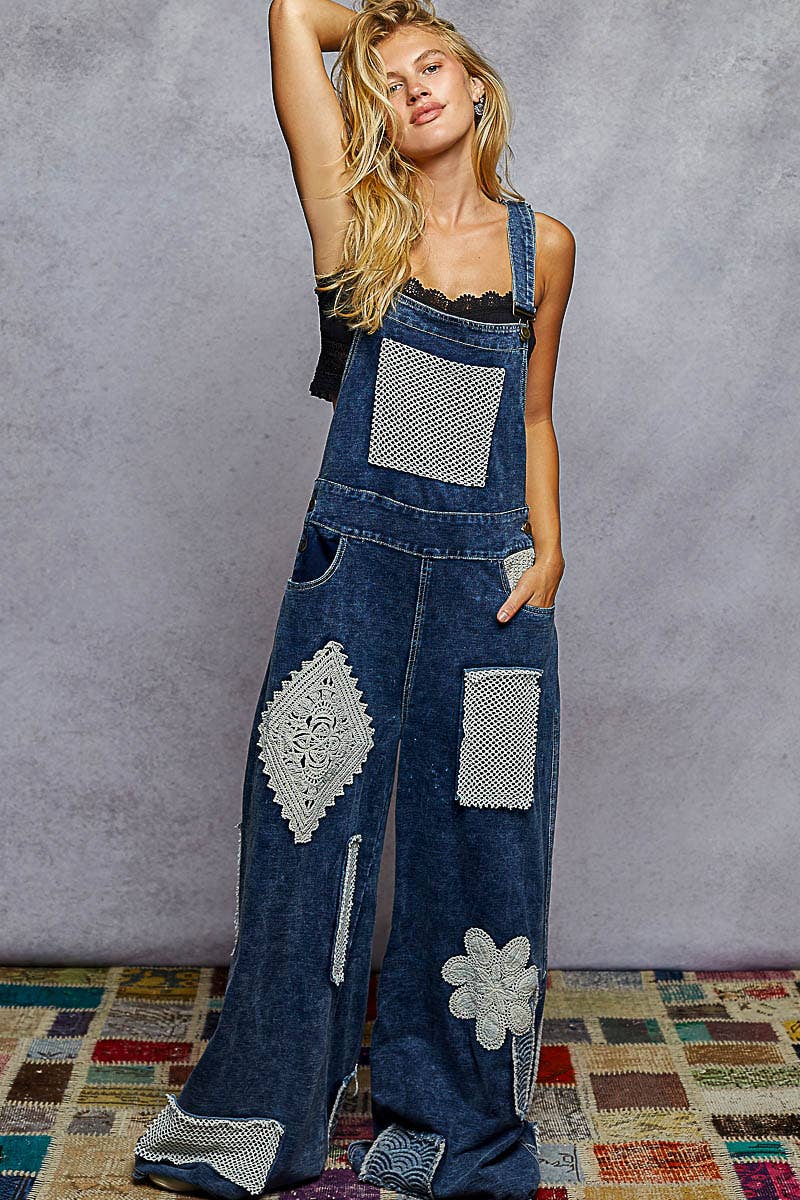 Boho denim patchwork overalls featuring wide legs, crochet lace appliqué patches, and vintage-inspired distressed detailing. Styled with a black crop top and sneakers for a relaxed, eclectic look.