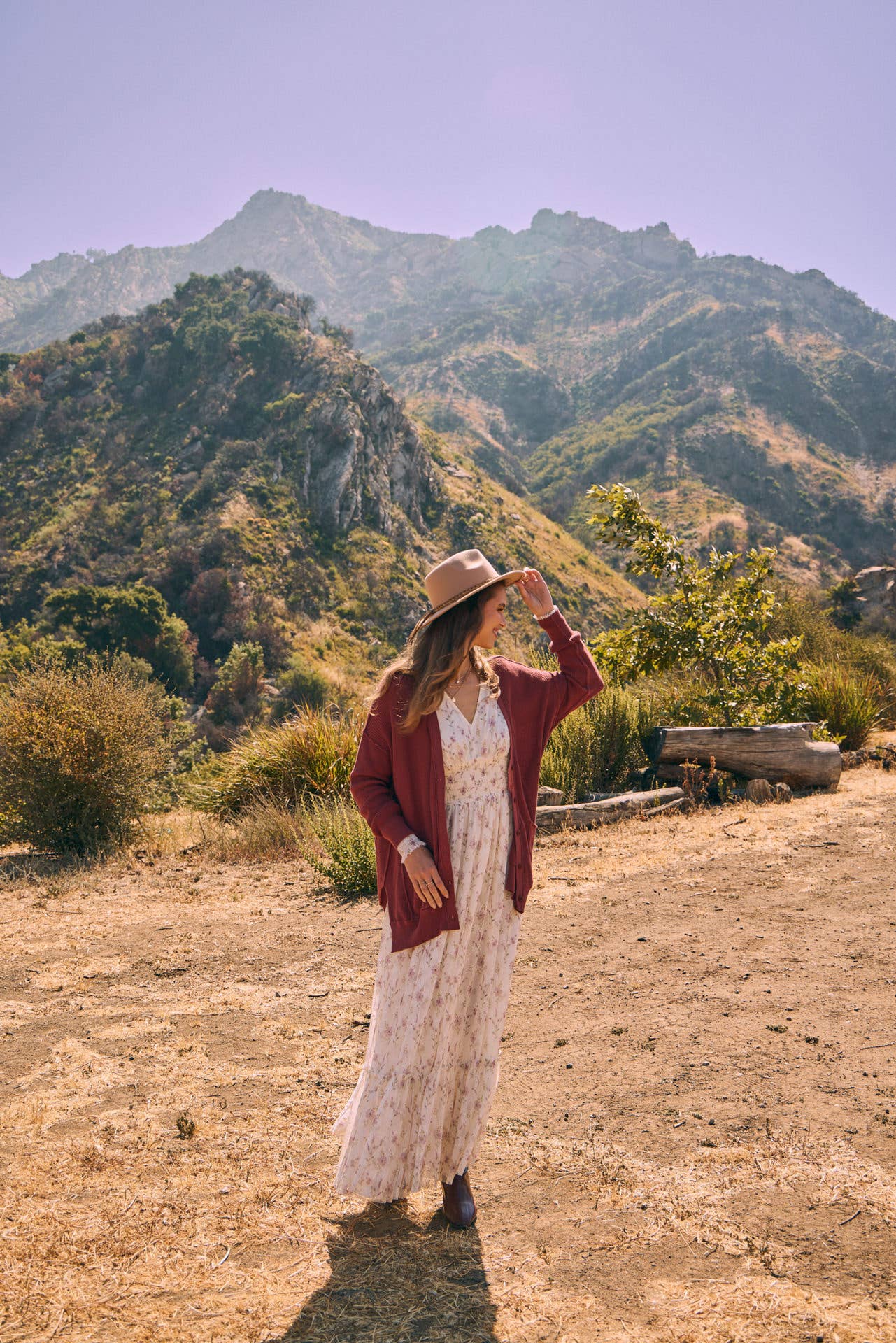 Front view — cream floral maxi dress with smocked waist and ruffle hem; mauve cardigan, tan hat, holding a greenery bouquet outdoors.