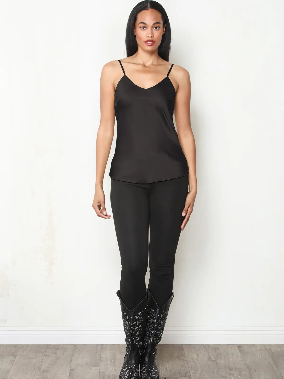 A woman modeling a black spaghetti-strap camisole top with black pants and black studded boots, standing against a white wall.