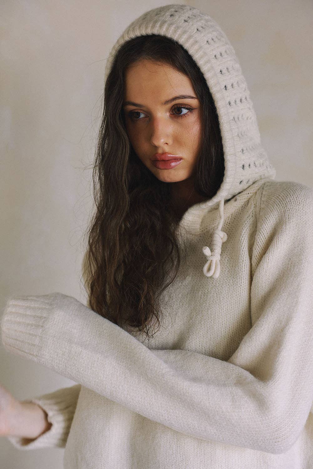 A woman wearing a cream-colored knit hooded sweater with a textured openwork hood and drawstrings, posed against a neutral beige background.