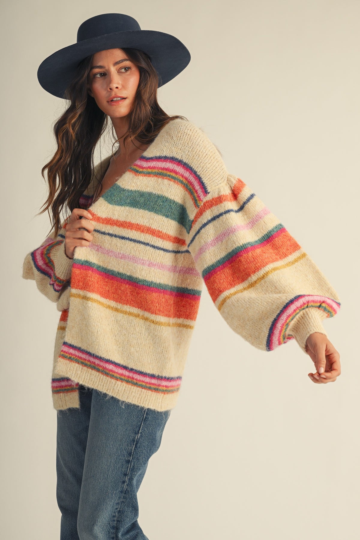 A woman wearing a cream-colored fuzzy knit sweater with vibrant multicolor horizontal stripes, paired with blue jeans and a black wide-brim hat, against a plain beige background.