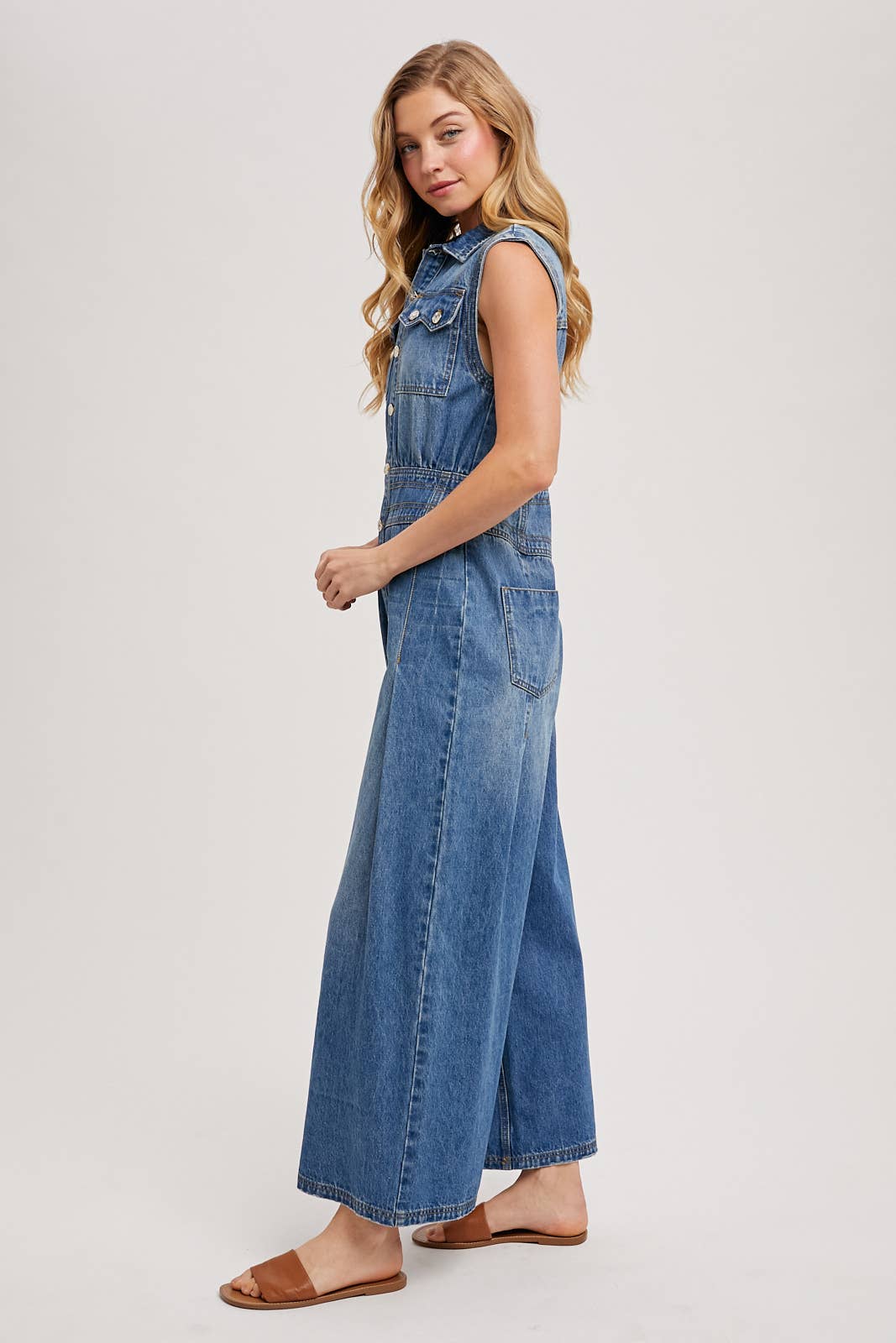 Side view of sleeveless denim jumpsuit with collared button-front and wide-leg legs, worn with brown slide sandals.