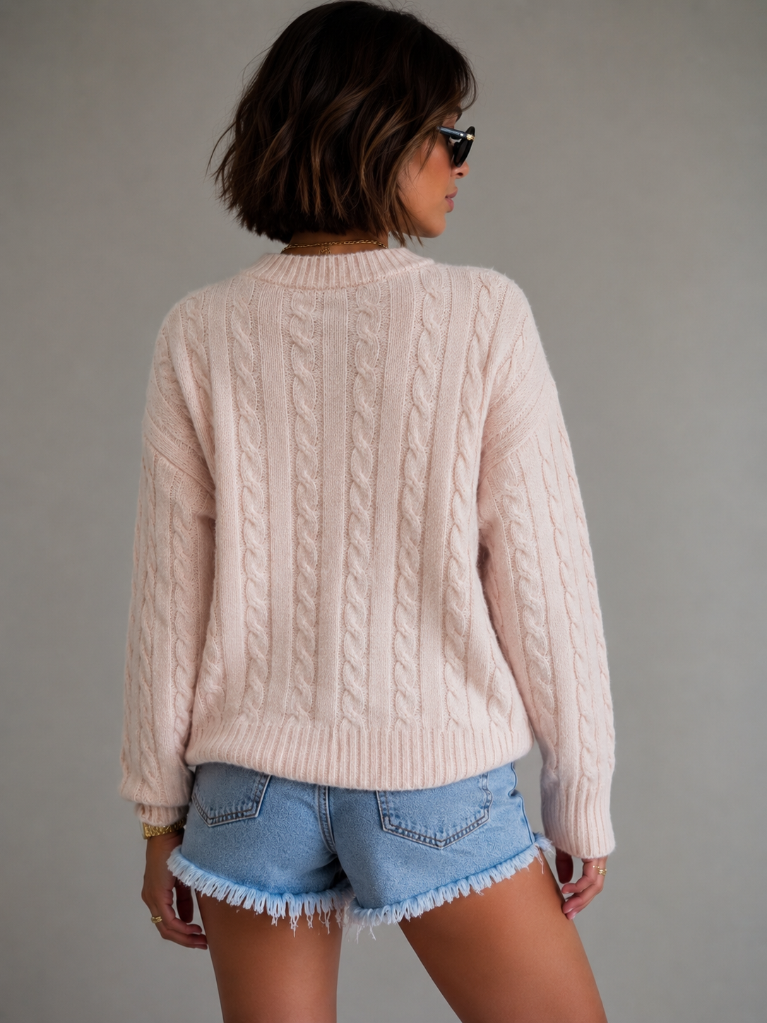 A woman shown from the back wearing a pale blush pink cable-knit sweater and light blue denim shorts against a plain gray studio background.