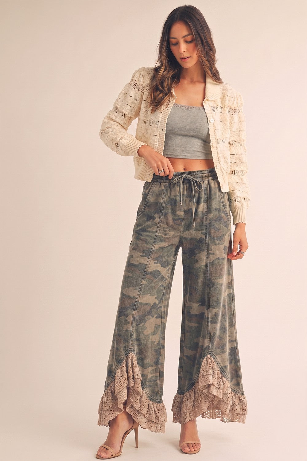 Woman wearing a beige jacket, gray top, and camouflage pants with ruffled hem on a plain background