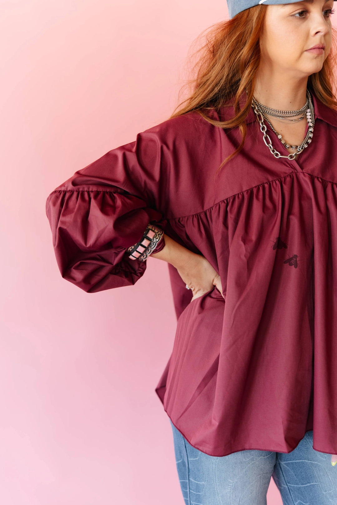 A woman wearing a loose-fitting, long-sleeve burgundy blouse with a gathered ruffle neckline and pockets, paired with blue jeans and layered silver necklaces, set against a solid pink background.